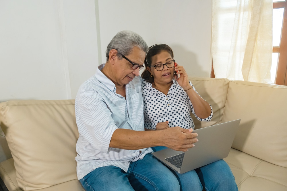 Latino senior couple applying for Medicare sitting on a couch, looking at a laptop, with one person talking on the phone.