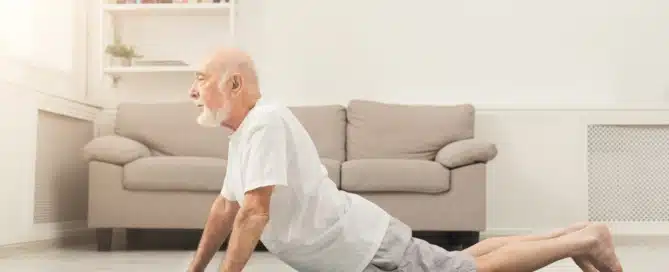 Senior man performing lower back stretching exercise on a yoga mat in a bright living room.