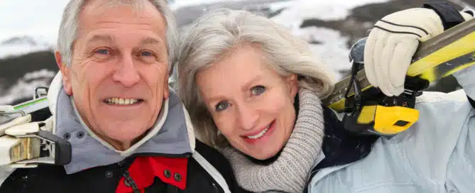 An elderly couple enjoying a day of skiing and winter outdoor exercise in a snowy mountainous landscape, dressed in winter gear and smiling.