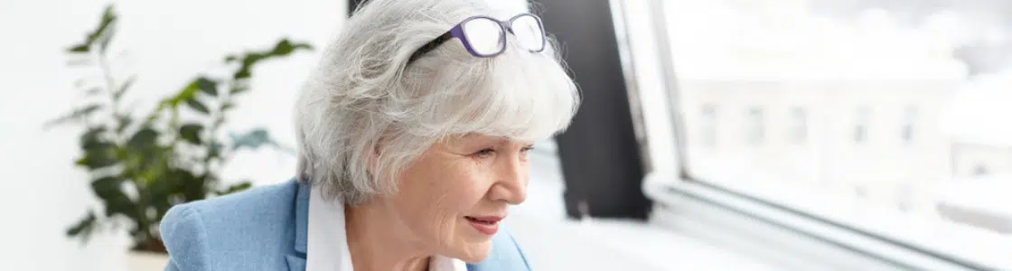 Senior woman in a light blue blazer working on a laptop at a desk, reviewing documents, representing the choice seniors face between employer insurance and Medicare at 65.