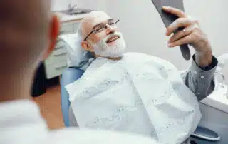 Senior man smiling in a dental chair while holding a mirror, reflecting satisfaction after a dental visit.