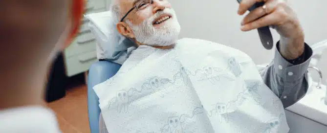 Senior man smiling in a dental chair while holding a mirror, reflecting satisfaction after a dental visit.