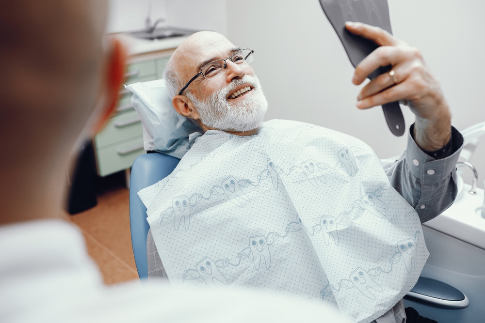 Senior man smiling in a dental chair while holding a mirror, reflecting satisfaction after a dental visit.